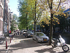 Typical gracht (city canal) with berthed boats on the water, cars on the embankments and a small bridge in the distance - Amsterdam, Hollanda